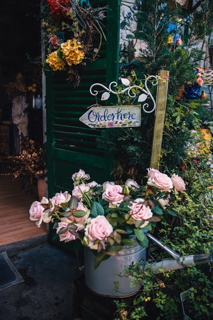 A display of fresh flowers and floral arrangements outside Leytonstone Florist, featuring a bouquet of pale pink roses with lush green foliage in a silver watering can, positioned on the ground near a