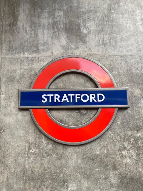 A close-up of a London Underground station sign displaying 'STRATFORD' in bold white letters on a blue background, mounted on a circular red and silver frame. The sign is attached to a textured grey c
