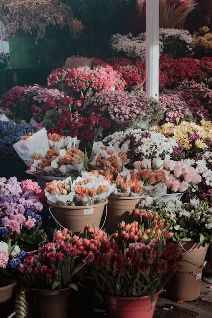 A vibrant display of fresh flowers arranged in various pots and buckets at a flower market, featuring an assortment of tulips, carnations, and chrysanthemums in colors including red, pink, white, yell