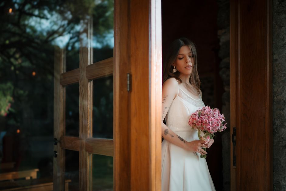 A young woman with shoulder-length brown hair and light skin, wearing a white sleeveless dress with lace detailing, stands partially behind a wooden door, holding a bouquet of pink hydrangeas. The bou