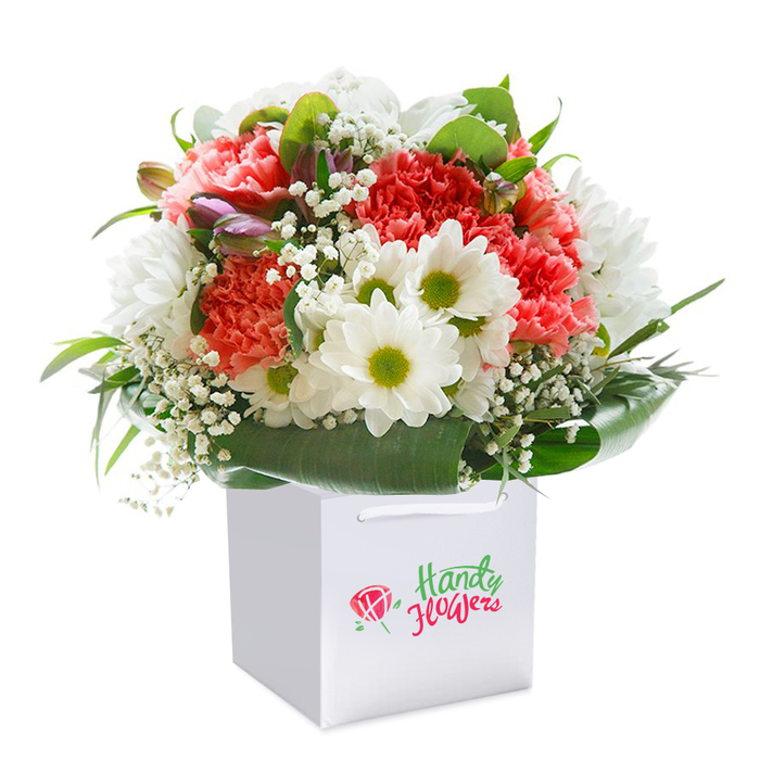 Perfectly poised within a flawless white gift bag, this lively bouquet showcases the very essence of Leytonstone's community spirit. Vibrant coral and peach carnations open at the bouquet's heart, their layered ruffles offering a plush, textural depth that feels both joyful and inviting-echoing celebratory moments in the E11 neighbourhood. White daisy-like blooms, each crowned with a zesty lime-green center, are strategically positioned for brightness, their simple charm reminiscent of morning walks in Bushwood's leafy parks. Speckles of airy white baby's breath thread delicately throughout, providing softness and light. Complementing these, a handful of purple-tinted floral buds peek from the left, adding intrigue and a sense of anticipation. The deep green foliage encircling the arrangement glistens, forming a lush, glossy frame that underscores the bouquet's freshness and expert handling. Every flower is artfully balanced for fullness, presenting an abundant and harmonious composition. The clean white gift bag, paired with matching ribbon handles, is designed for seamless gifting across Leytonstone, whether for anniversaries, birthdays, or spontaneous gestures. All focus remains on the artistry-no logos visible-making the image ideal for illustrating expert local floristry, the joy of flower delivery, or inspiration for thoughtful gifts within the Leytonstone area. This bouquet, tenderly prepared by Leytonstone Florist, embodies the heart and creativity of its vibrant London community.