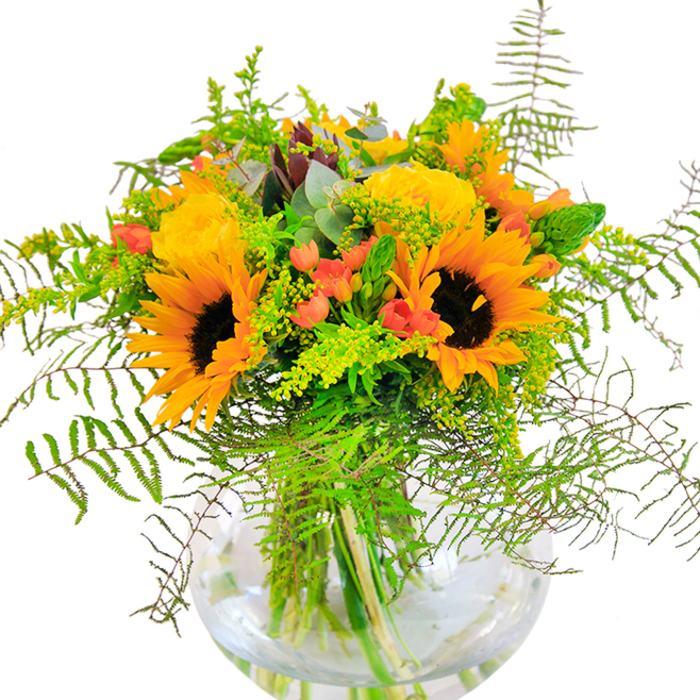 A first look at this bouquet instantly conjures images of sunlit summer days in Leytonstone, where sunflowers and yellow roses beam side by side, their layered petals unfurling with exuberant warmth. Deep green foliage and airy, cascading vines give the arrangement its lush, garden-like character, reminiscent of the vibrant displays at Leytonstone Farmers Market. Scarlet Hypericum berries glint among the leafage, their shiny surfaces contrasting with the soft, velvety petals and the matte leaves. Everything is gathered into a rounded glass vase, its clarity revealing the artful architecture of stems nestled in fresh water. Moving closer, the bouquet's rich textures are felt: the suede disks of sunflowers, the tightly wound rose buds, the sprightly snap of greenery. The fragrance is gentle, a blend of zesty greens and mellow blossoms that stirs echoes of nature's vitality. As a thoughtful gift for a milestone birthday, a congratulatory bouquet for a new neighbour on Church Lane, or a centrepiece for a cozy dinner in E11, this arrangement channels the best of Leytonstone-joyful, harmonious, and alive with the colours of the season.